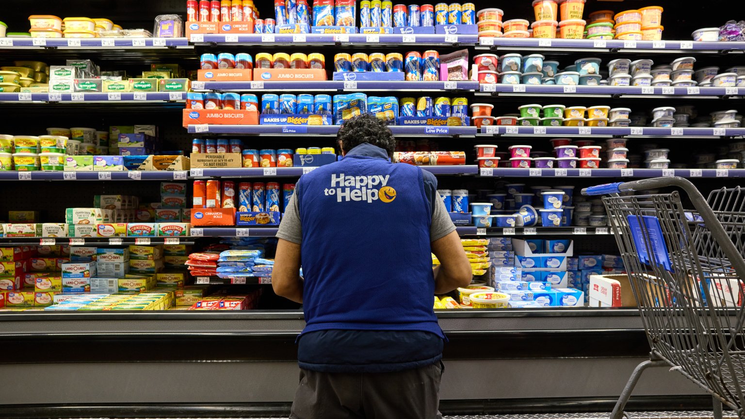 An employee in a blue vest kneels in front of a refrigerated section of a grocery store.