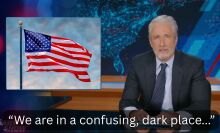 A man in a suit sits behind a talk show desk, with an image of the American flag in the top-left.