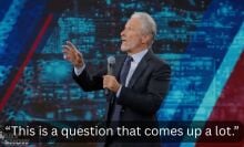 A man stands on a stage in a suit with a microphone, pointing out towards the audience.