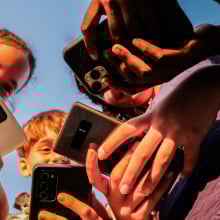 A group of young kids smile and hold out several different models of phones while huddled in a group.