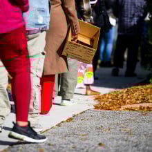 People stand in line holding cardboard boxes and grocery bags. 