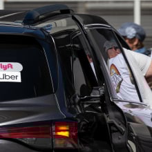 A person gets into the back of a dark car with stickers advertising Uber and Lyft. 