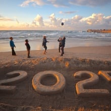 Children on a beach playing with a ball by a '2025' sand sculpture