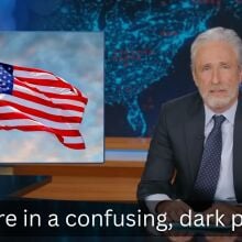 A man in a suit sits behind a talk show desk, with an image of the American flag in the top-left.