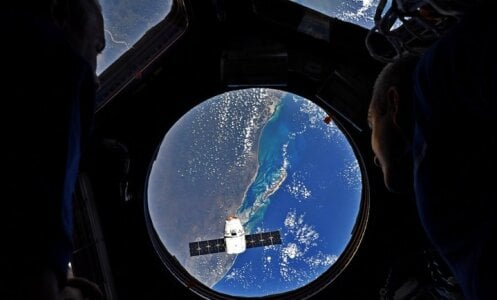Astronauts in the ISS look out through a window to see the SpaceX Dragon vessel floating over the Earth. The Drago carried OCO-3 to the ISS.