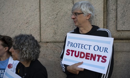 A person stands in front of a stone building holding a sign that reads "protect our students."