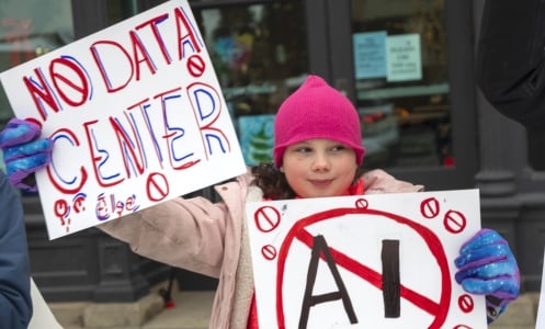 A young anti-AI protestor in a pink hat holds "No Data Center" and "No AI" signs