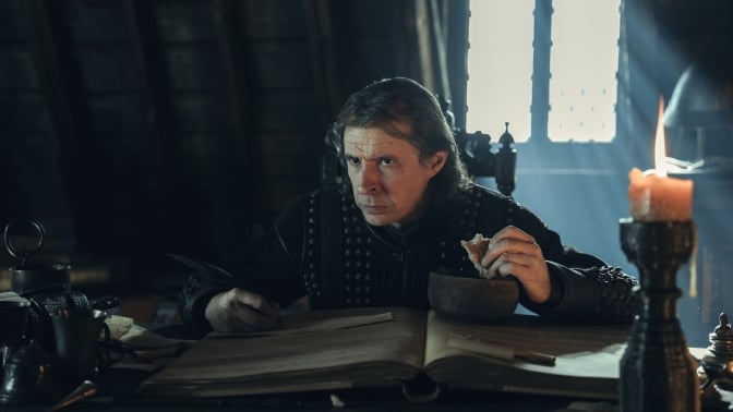 A man sits at a desk in a dark room, eating while he works.