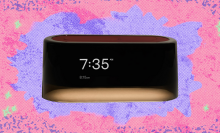 A rectangular, modern alarm clock with a black display showing "7:35 AM" and "8:15 AM" in white text, set against a vibrant, abstract background of pinks, purples, and blues with a splattered, textured effect.
