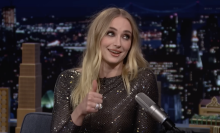 A woman sits on a talk show sofa grinning and holding up her thumb.