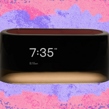 A rectangular, modern alarm clock with a black display showing "7:35 AM" and "8:15 AM" in white text, set against a vibrant, abstract background of pinks, purples, and blues with a splattered, textured effect.