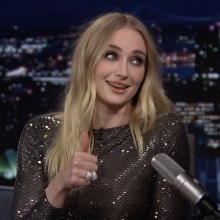 A woman sits on a talk show sofa grinning and holding up her thumb.