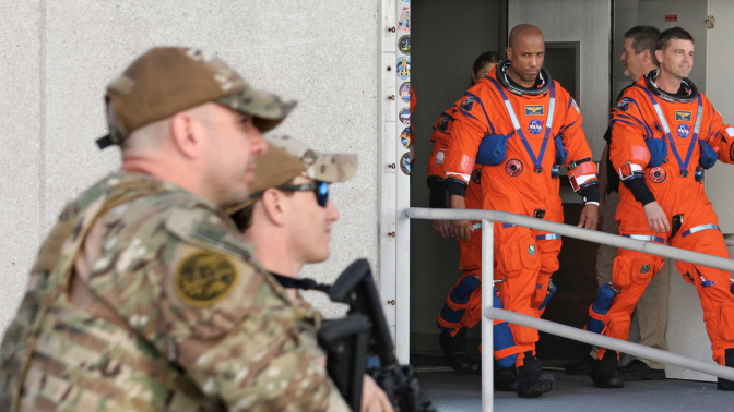 A group of Artemis II astronauts in orange suits walk out of a building guarded by armed soldiers. 